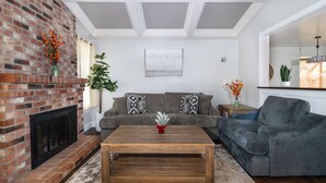 A living room with a brick fireplace, a gray sectional sofa, a wooden coffee table, and a large window with curtains. - Comfy House W/ Big Pool (Elk Grove)