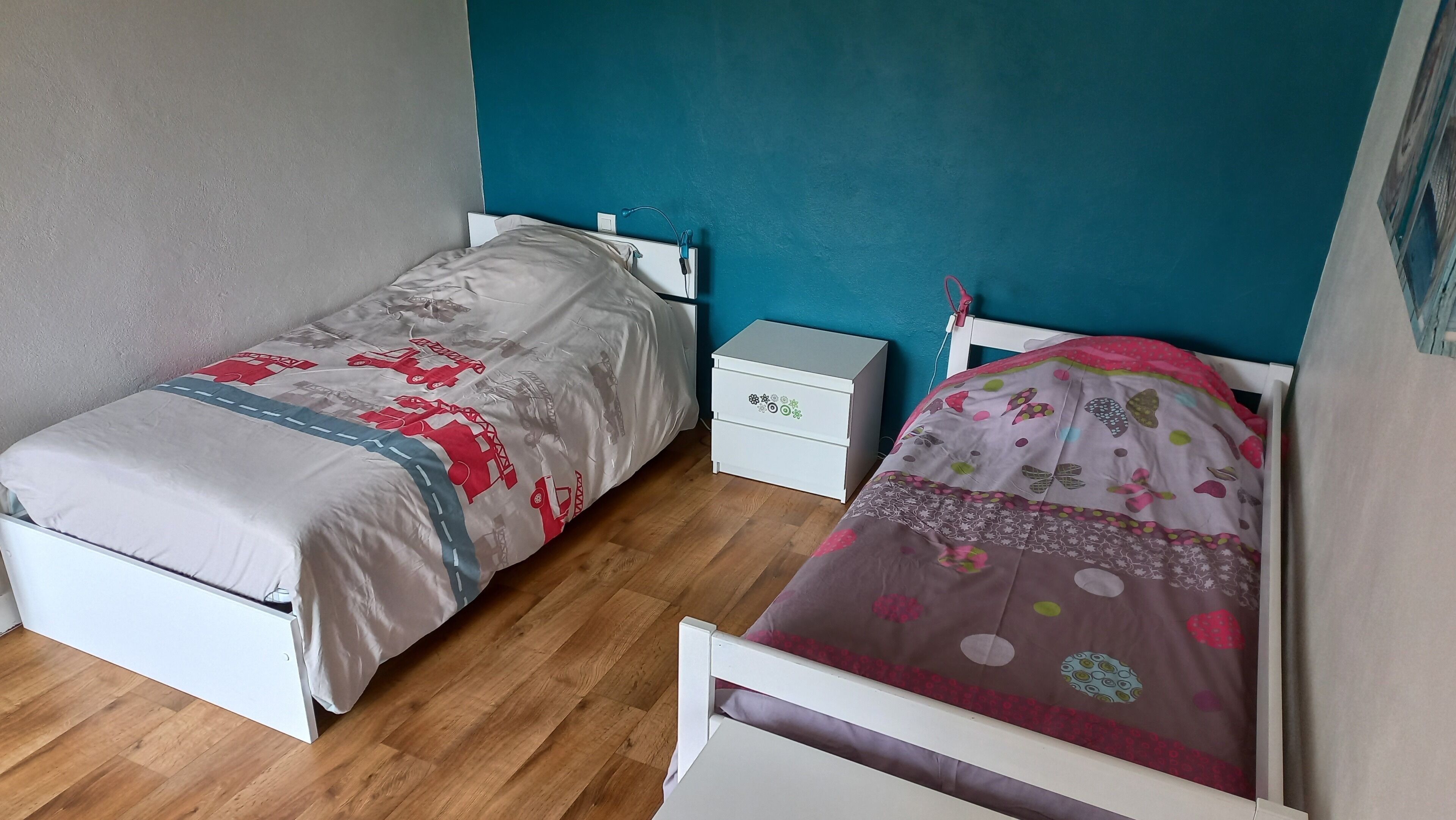 A room with two single beds, a white nightstand with drawers, and a teal accent wall.