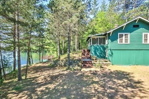A green cabin with a stone fireplace, surrounded by trees and a lake.