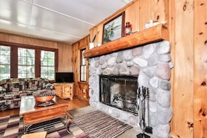A cozy living room with a stone fireplace, wooden walls, a patterned sofa, and a television.