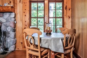 Ein Holzblockhaus-Interieur mit Kamin aus Stein, einem runden Esstisch und einem großen Fenster mit Blick auf Bäume.