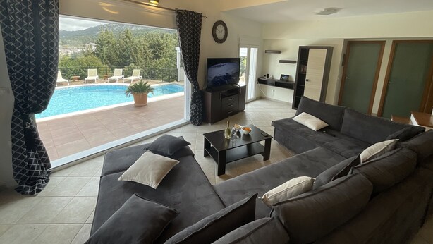 A spacious living room with a large sofa, a TV, and a view of a swimming pool. - villa aphrodite (Maritsa)