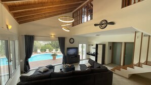 A spacious living room with a large sectional sofa, a television, and a view of a swimming pool. - villa aphrodite (Maritsa)