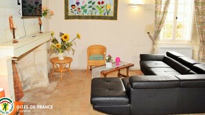 A living room with a black leather sofa, a wicker chair, a small round table with a vase of sunflowers, and a framed painting of flowers and birds on the wall. - Spacious Historic Presbytery, Sleeps 15, Garden, WiFi, Near Châteaux & La Roche-Posay (Bossay-sur-Claise)