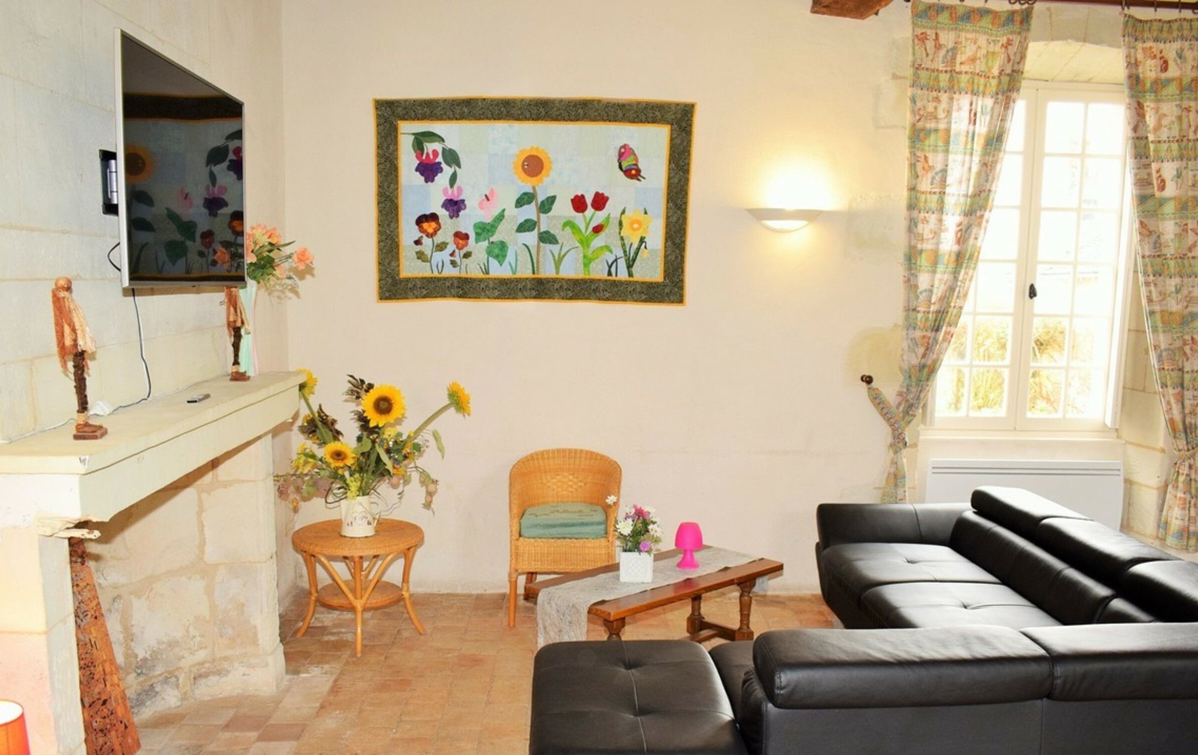 Un salon avec un canapé en cuir noir, un fauteuil en osier, une petite table ronde ornée d’un vase de tournesols et, au mur, un tableau encadré représentant des fleurs et des oiseaux.