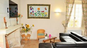 Een woonkamer met een zwarte leren bank, een rieten stoel, een klein rond tafeltje met een vaas vol zonnebloemen en een schilderij van bloemen en vogels aan de muur.