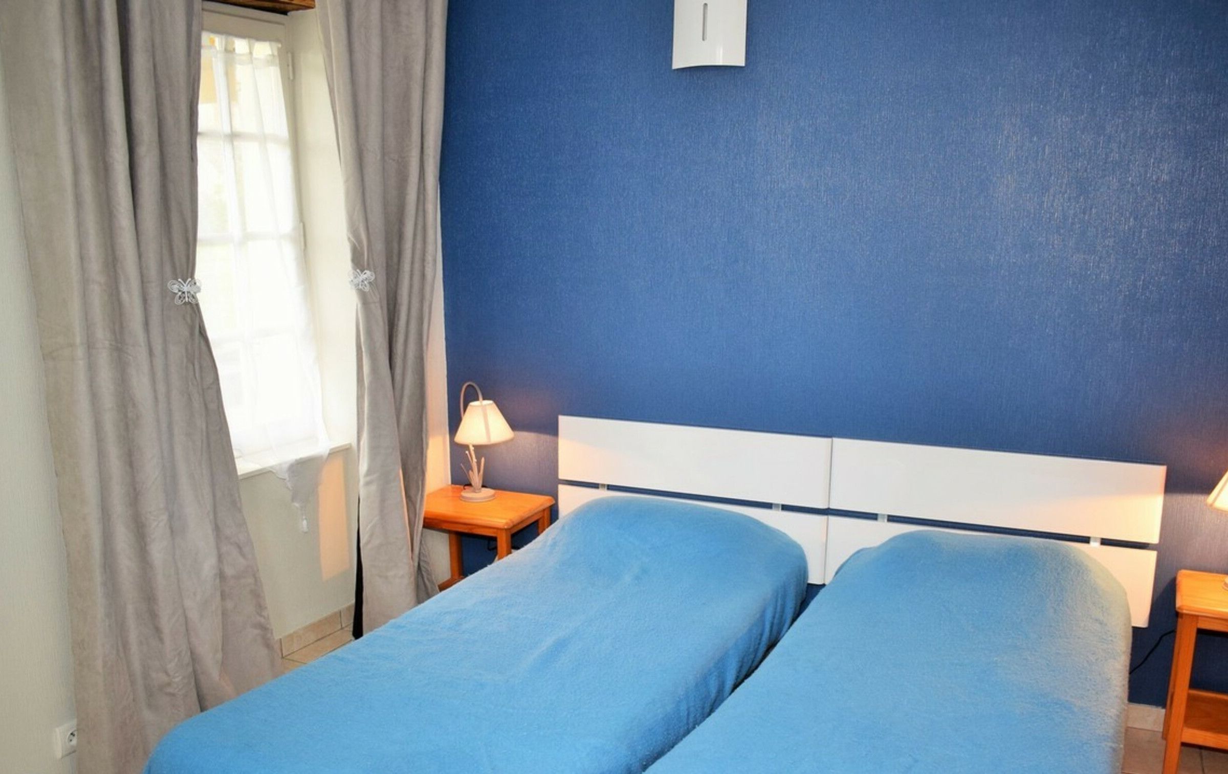 A room with two single beds, a nightstand with a lamp, a window with curtains, and a blue wall.