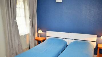A room with two single beds, a nightstand with a lamp, a window with curtains, and a blue wall.