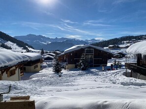 A snow-covered mountain village with wooden buildings, a central lodge, and a clear view of the surrounding mountains.