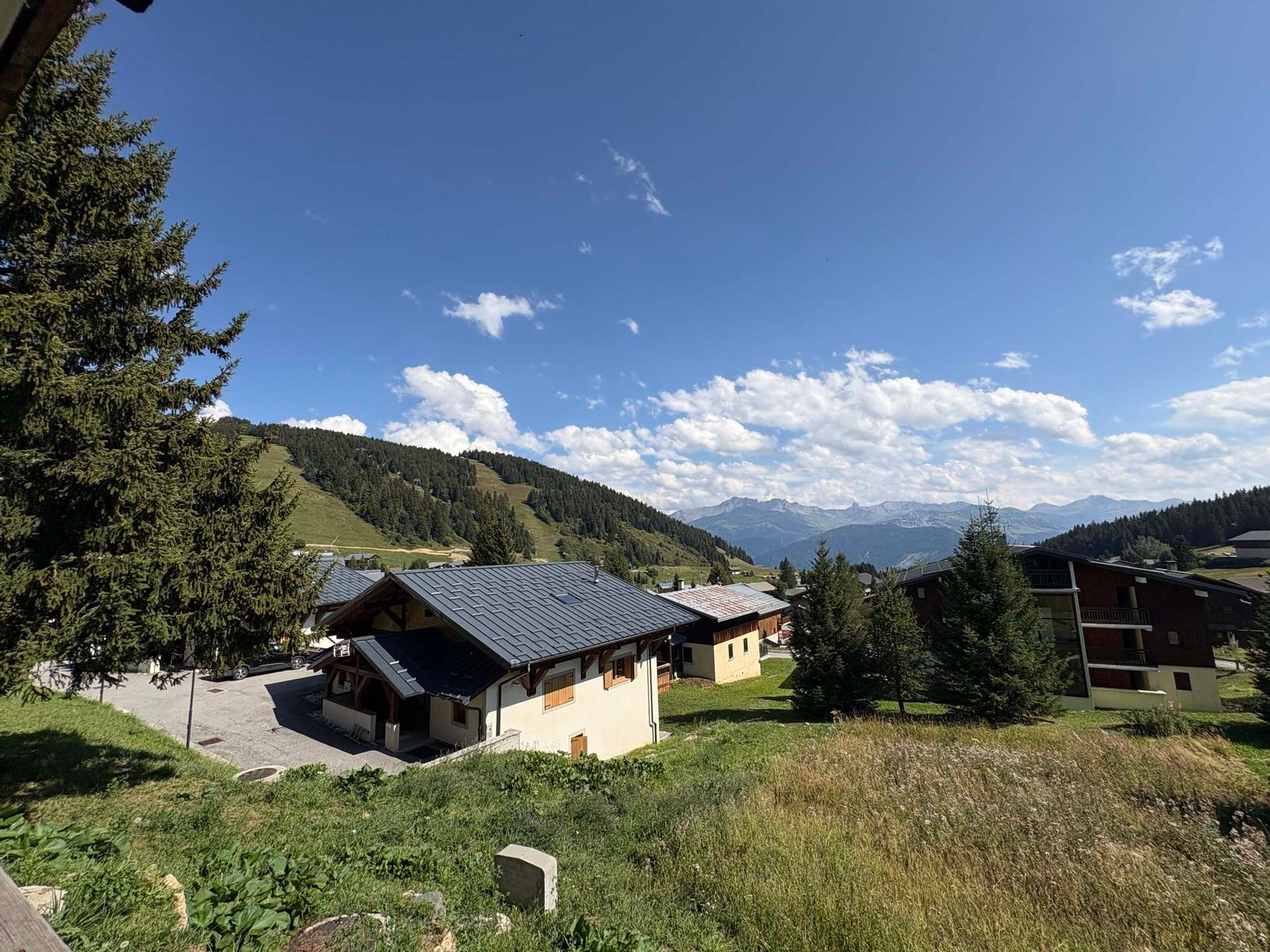 A mountain cabin with a sloped roof, surrounded by greenery and a view of distant mountains.