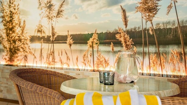 A wicker chair with a yellow and white striped cushion, a glass pitcher with ice, and a glass on a round table with a view of tall grasses and a body of water. - Holiday Apartment "Alpenhof Lodge Ifinger" with Mountain View, Breakfast, Wellness & Wi-Fi (Saltaus)