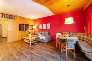 Une pièce chaleureuse avec un plafond en bois, un mur d’accent rouge, un canapé, une table à manger avec des chaises et une petite table sur laquelle est posé un vase.