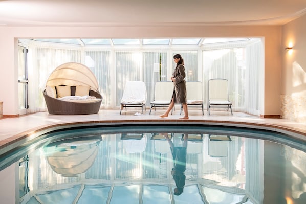 Un espace piscine intérieur moderne, doté d’un coin salon fermé par des baies vitrées, où l’on aperçoit une personne qui se promène.