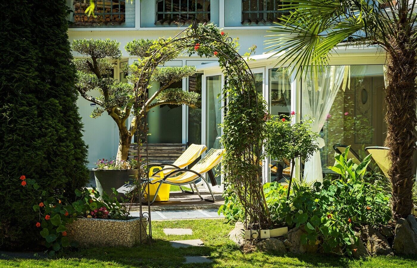 A well-maintained garden with a variety of plants, a seating area with yellow chairs, and a glass-enclosed structure.
