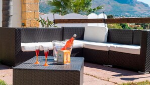Un canapé en osier avec un coussin blanc, une petite table avec deux verres et une bouteille, et une vue sur des montagnes en arrière-plan.