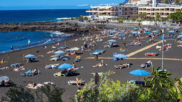 Una playa con numerosas tumbonas y sombrillas, una costa rocosa y edificios al fondo.