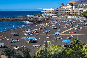 A beach with numerous sun loungers and umbrellas, a rocky shoreline, and buildings in the background.