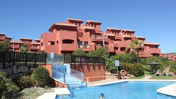 A swimming pool with a slide, surrounded by a red building with balconies, and a clear blue sky. - Apartment 'Duplex Estepona' with Mountain View, Wi-Fi and Air Conditioning (Estepona)
