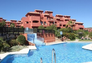 A swimming pool with a slide, surrounded by a red building with balconies, and a clear blue sky.