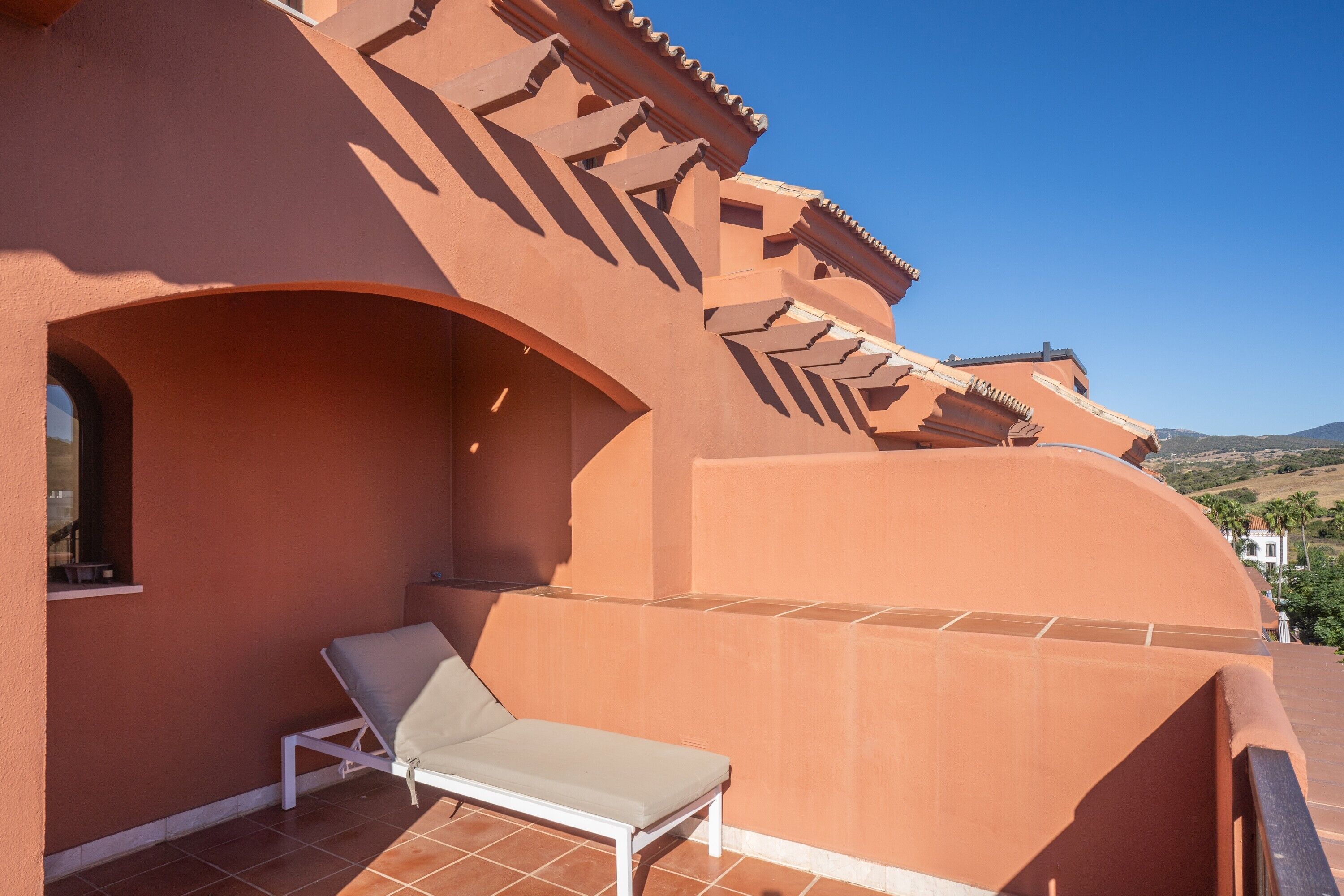 A sunlit balcony with a single lounge chair, a tiled floor, and a clear view of the surrounding landscape.