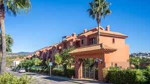 A row of orange buildings with a gated entrance, palm trees, and a clear blue sky. - Apartment 'Duplex Estepona' with Mountain View, Wi-Fi and Air Conditioning (Estepona)