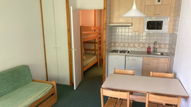 A compact room with a bunk bed, a small kitchenette with a microwave and stove, a dining table with chairs, and a green sofa.