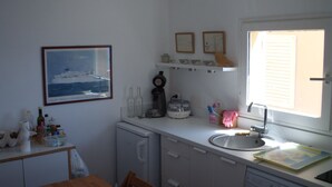 A compact kitchen with a small dining table, a refrigerator, a coffee maker, and a window with blinds. - Ile du Frioul, 4-bed apartment at the foot of the calanques (Marseille)