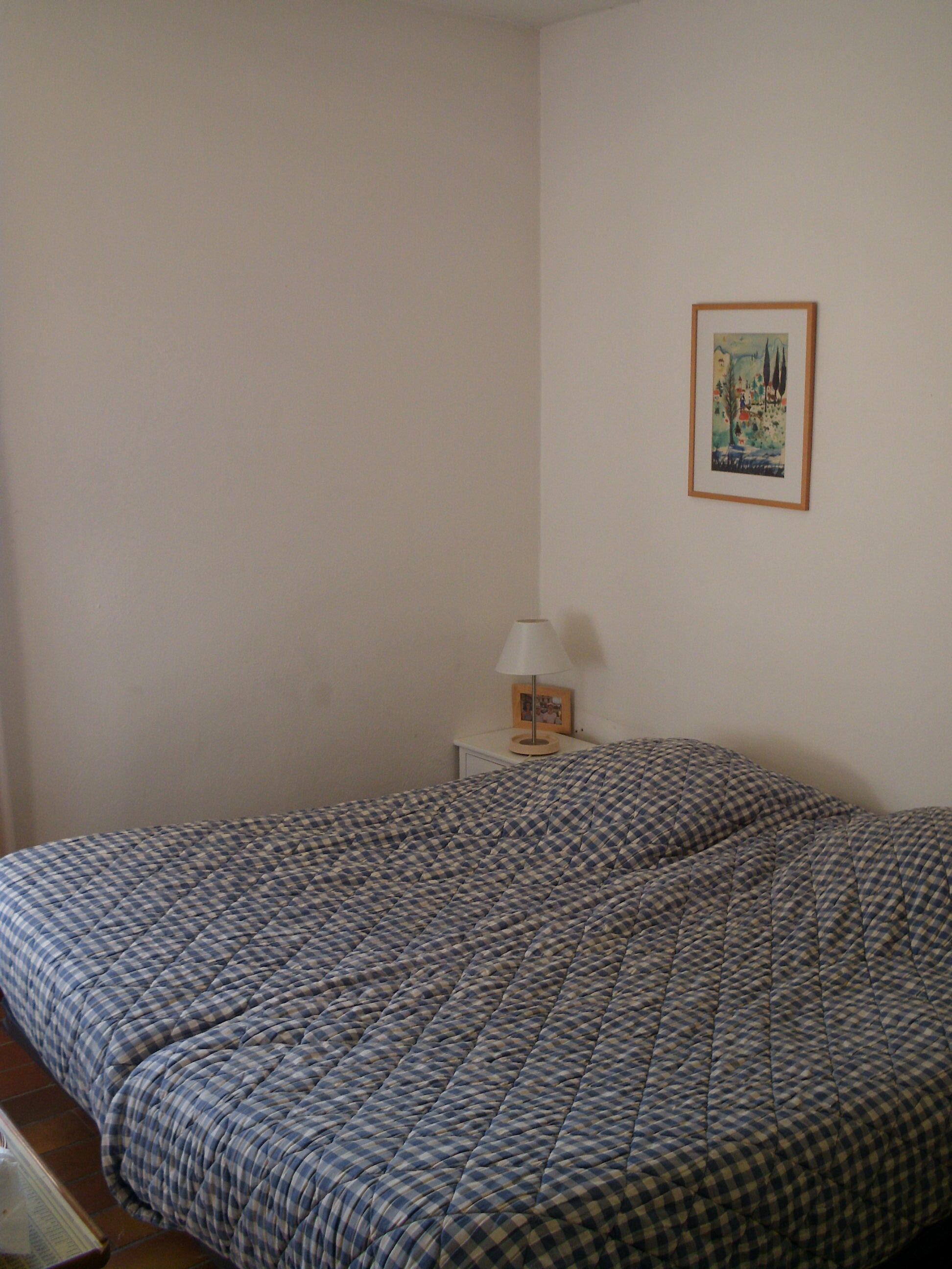 Ein Schlafzimmer mit kariertem Bettbezug, einem Nachttisch mit Lampe und einem gerahmten Bild an der Wand.