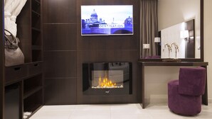 A modern hotel room with a TV displaying a cityscape, a fireplace, a vanity with a mirror, and a purple chair. - Roman Holidays Boutique (Rome)