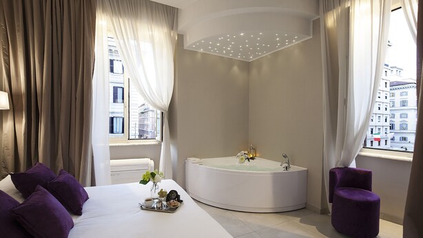 A modern hotel room with a large bed, a round bathtub, and a small purple chair. - Roman Holidays Boutique (Rome)