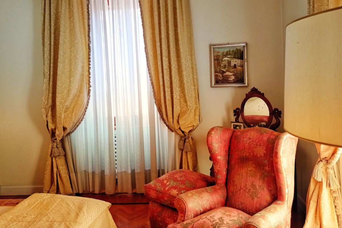 A room with a large window, a red armchair, a wooden chair, a mirror, and a bed with a quilt.
