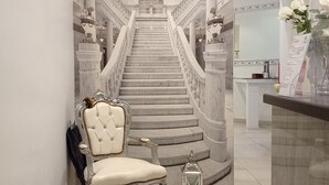 A grand staircase with a marble finish, leading to a balcony with a view. A plush chair and a small lantern are placed near the stairs. - Roman Holidays Boutique (Rome)