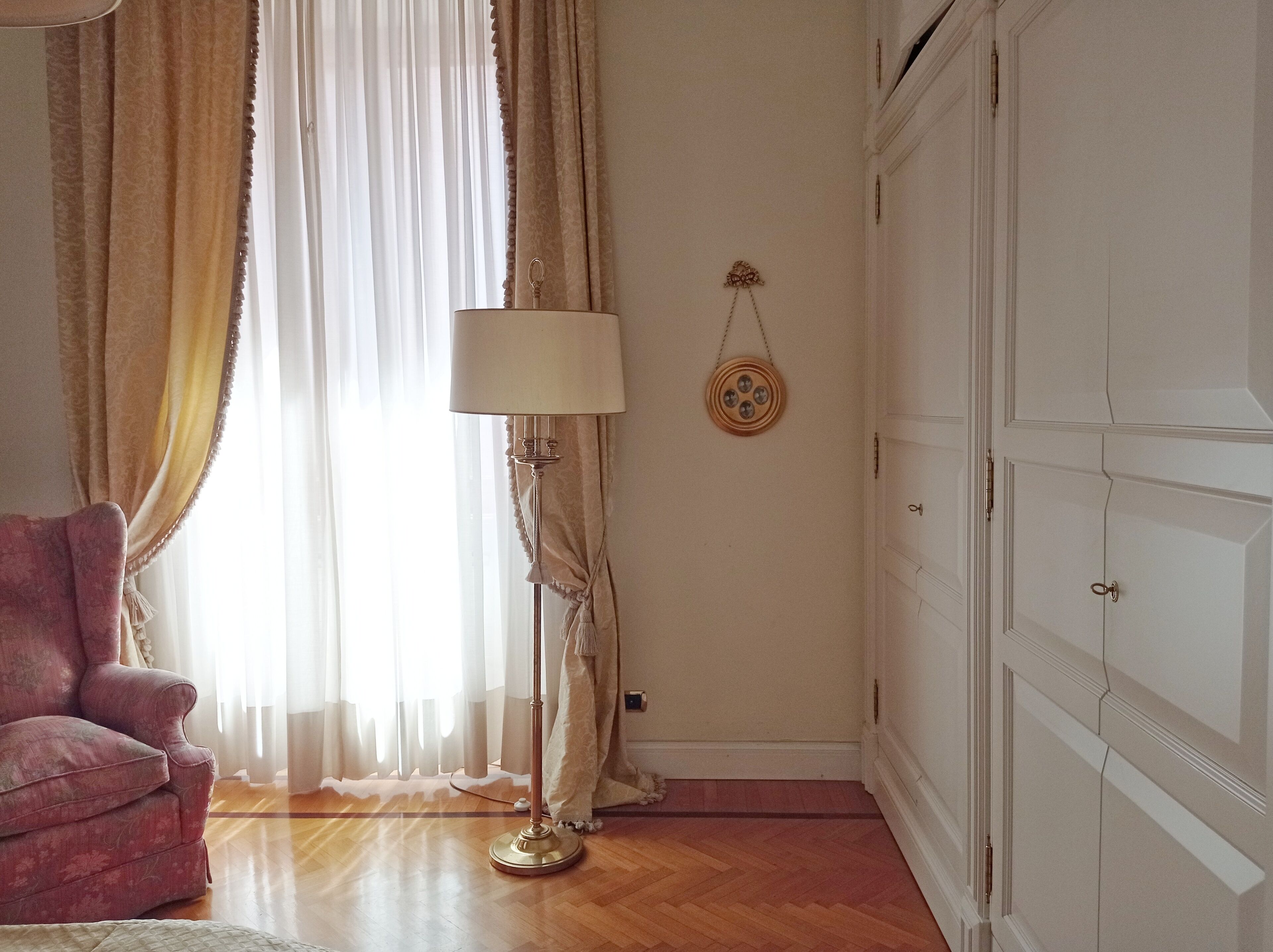 A room with a pink armchair, a floor lamp, a wardrobe, and a window with sheer curtains.