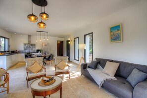 A spacious living area with a grey sofa, two armchairs, a coffee table, and pendant lighting. A kitchen is visible in the background with a dining table set for four.