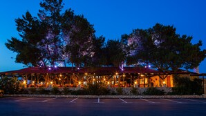 A covered outdoor dining area with multiple tables and chairs, surrounded by trees and a stone wall. - Hostel Vagabundo (Novalja)