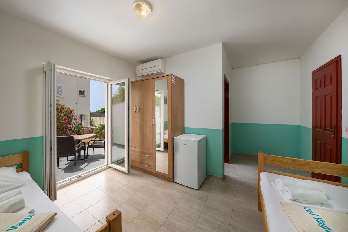 A room with a bed, a small refrigerator, a wooden wardrobe, and a sliding glass door leading to an outdoor area with a table and chairs.