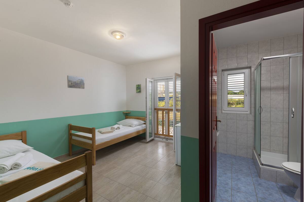 A room with two wooden beds, a bathroom with a shower, and a view through a door.