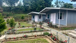 A two-story building with a flat roof and a balcony. There are flower beds with red flowers in front of the building. - KedaraYatih Resort (Ukhimath)