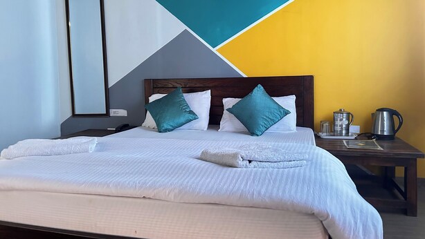 A neatly made bed with white linens and teal pillows, a bedside table with a kettle and glasses, and a geometric wall design. - KedaraYatih Resort (Ukhimath)
