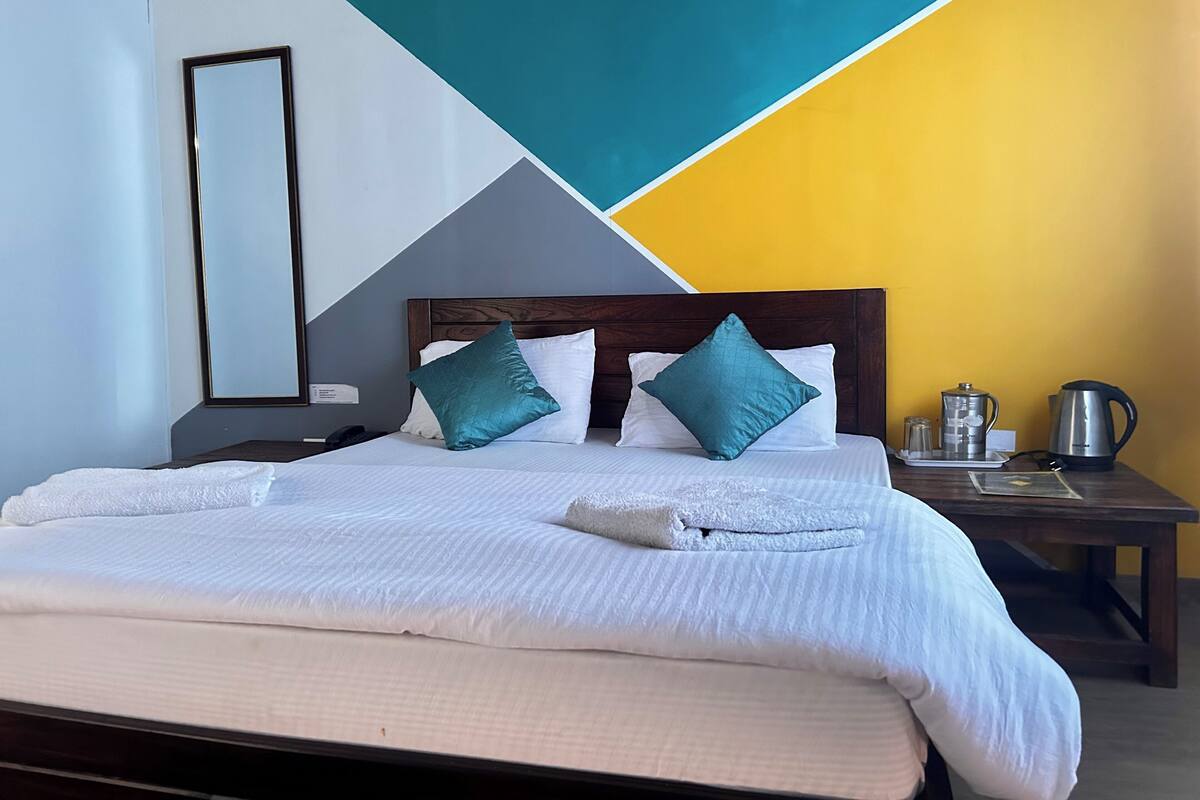 Un lit bien fait, avec du linge de lit blanc et des coussins turquoise, une table de chevet avec une bouilloire et des verres, et un motif géométrique sur le mur.
