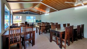 A spacious dining area with wooden tables and chairs, ceiling fans, and large windows offering a view of the outdoors. - KedaraYatih Resort (Ukhimath)