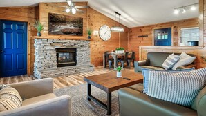 A cozy living room with a stone fireplace, wooden walls, a blue door, and a dining area with a table and chairs. - TopOfTheMountain, Fully Paved Roads, PoolTable, HotTub, VideoGames,EV Charger (Sevierville)