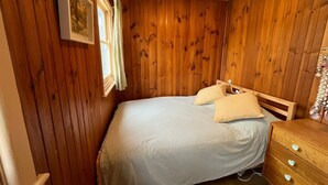 Una habitación con paneles de madera, una cama con colchón blanco y dos almohadas, un tocador de madera con cajones, y una lámpara colgante.