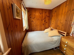 A room with wooden panel walls, a bed with a white mattress and two pillows, a wooden dresser with drawers, and a hanging light fixture.