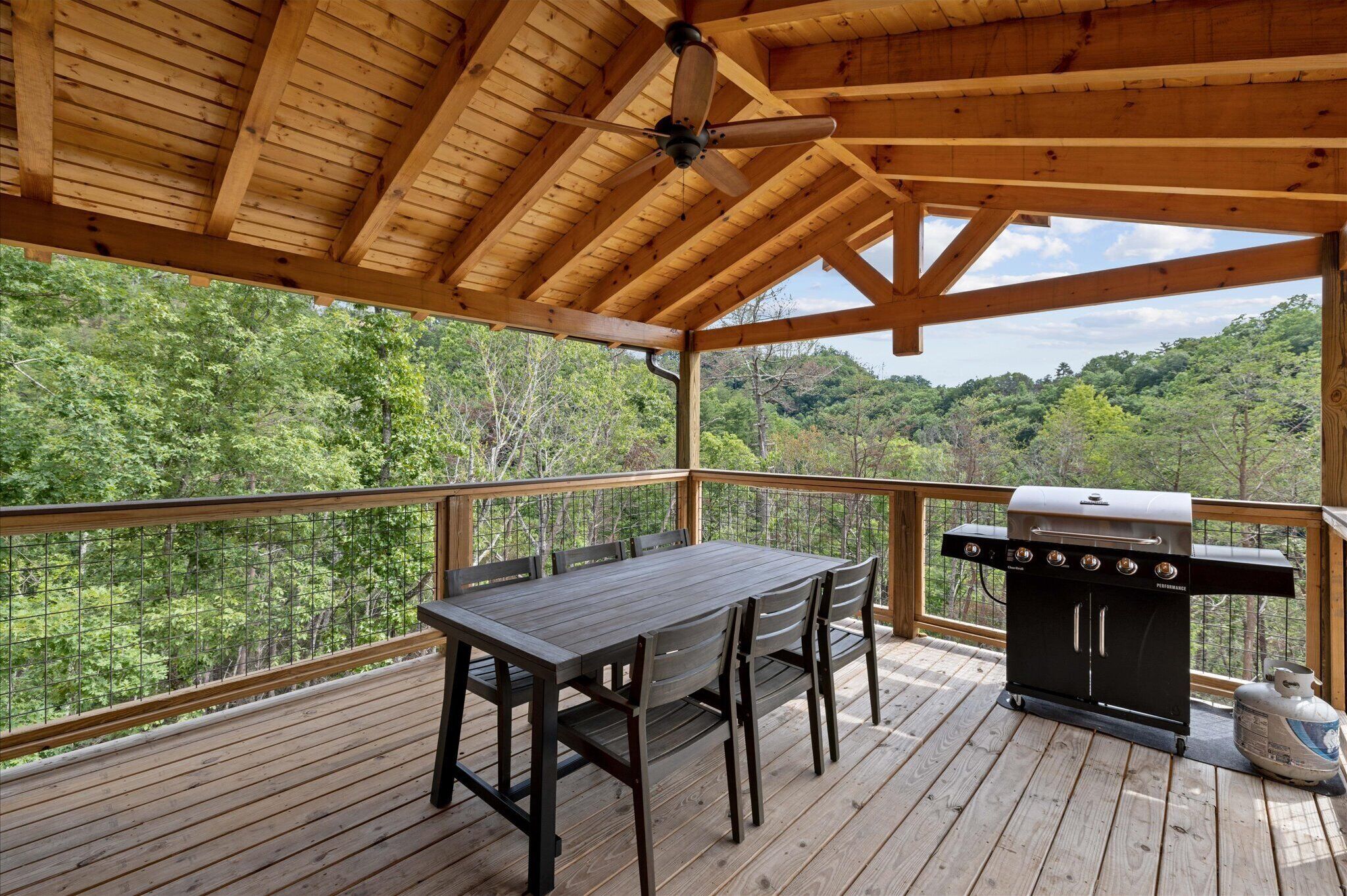 A covered deck with a wooden table and chairs, a grill, and a view of a forested area.