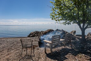A rocky shoreline with a wooden bench and a metal chair, a tree providing shade, and a body of water in the background. - Lake Front Retreat with boat access and private beach (Lowbanks)