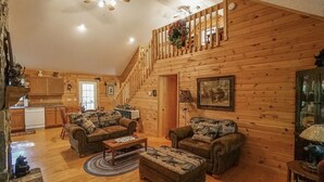 L’intérieur d’une cabane en rondins, avec un escalier en bois, deux canapés ornés de coussins à motifs animaliers, un coin repas et une cuisine.