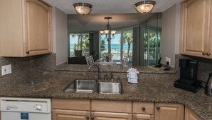 A kitchen with granite countertops, stainless steel sink, wooden cabinets, and a view of the ocean through large windows.
