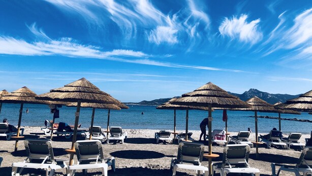 Una playa con sombrillas de paja y sillas de descanso, cielo azul despejado y mar tranquilo.