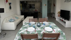 Una sala de estar moderna con comedor, sofás blancos, televisión y cocina al fondo.